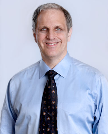 close up headshot of older man in button up shirt and tie smiling against baby blue wall